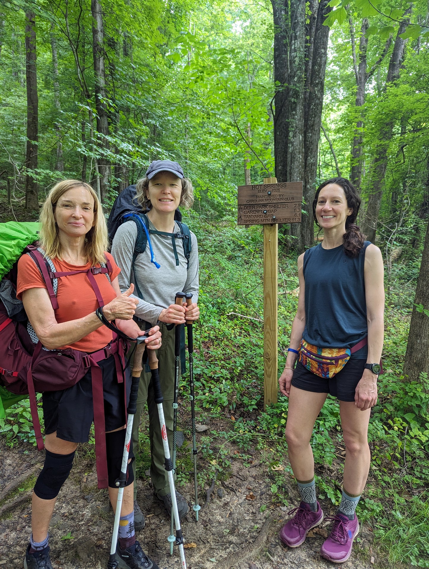 Three women with backpacks stand near a hiking trail sign
