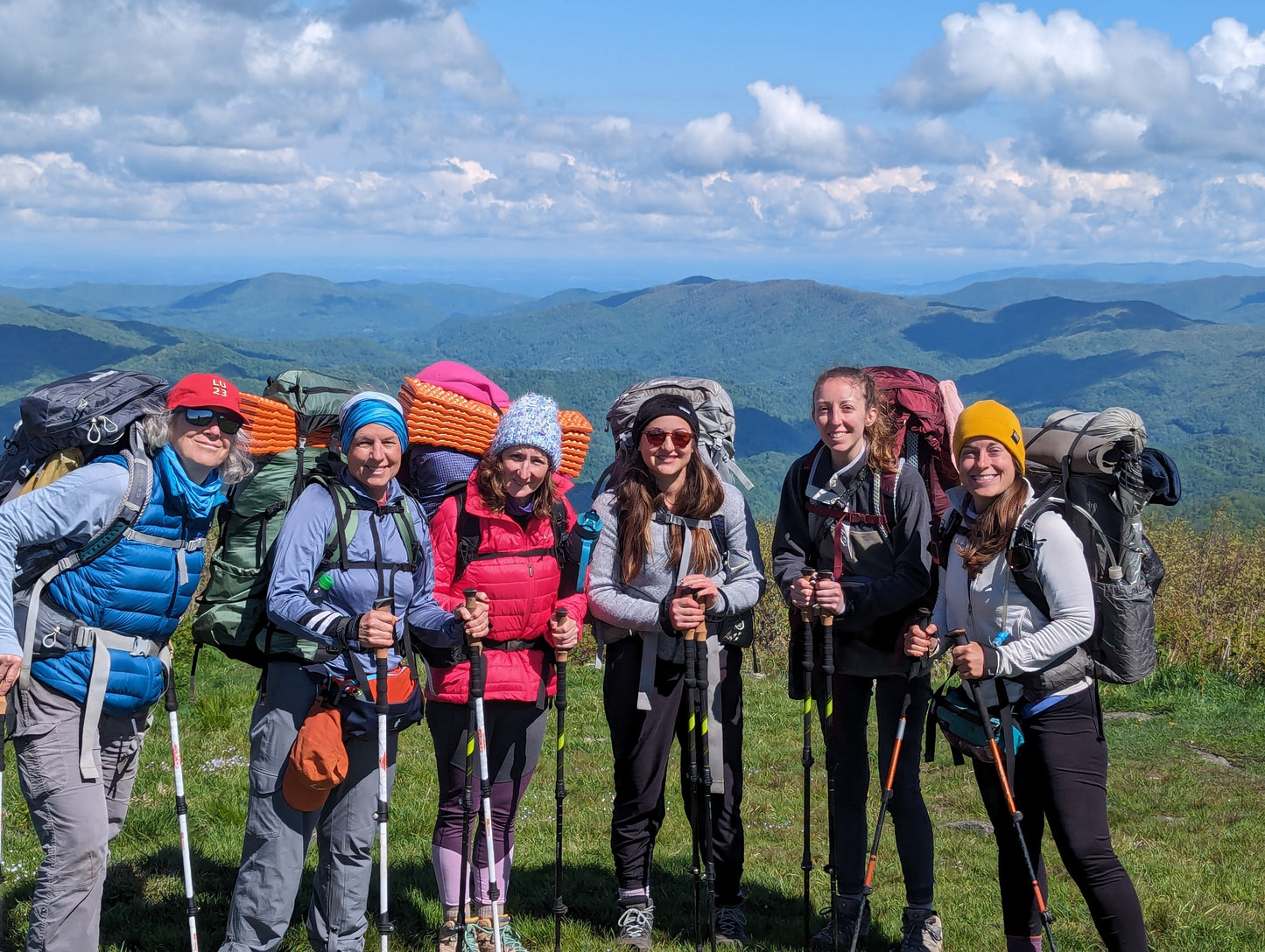 A group of six women standing on a mountain summit with backpacking gear and trekking poles